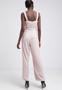 Vaaleanpunainen haalari, jossa on leveät lahkeet, muotoillut hihnat ja vyötäröllä vyö. Silkkinen kangas, joka istuu hyvin ja takana zip. Korkokengät.