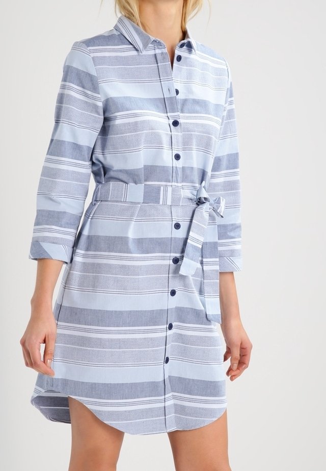 Femme portant une robe-chemise boutonnée à rayures horizontales bleu clair et blanc, avec une ceinture à la taille et des manches trois-quarts.