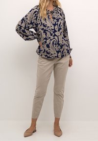 Blouse bleu marine avec motif cachemire beige, manches longues et détail froncé au col, associée à un pantalon ajusté beige clair et des ballerines nude.