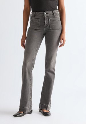 Žena odjevena u sive hlače visokog struka ravnih nogavica s prednjim džepovima i crnim šiljastim ravnim cipelama, stoji na jednobojnoj pozadini.