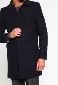 Cappotto di lana blu navy con colletto e chiusura a un bottone, dotato di tasche laterali e maniche lunghe, mostrato in vista frontale.