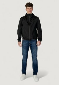 Giacca impermeabile nera con design aderente, zip frontale e cappuccio regolabile, abbinata a jeans blu e sneakers bianche.