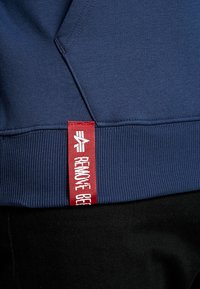 Felpa blu navy con texture a coste. Presenta un'etichetta rossa ben visibile con la scritta "REMOTE GEE" cucita all'orlo, in contrasto con il tessuto principale.
