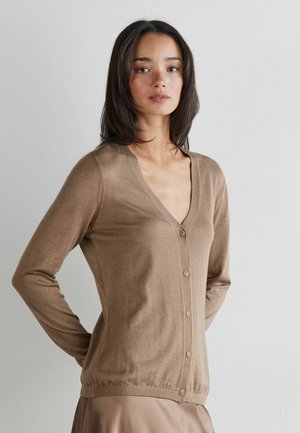 Vrouw draagt een lichtbruine gebreide cardigan met knopen en een bijpassende rok, staat met haar handen achter haar rug tegen een effen witte muur.