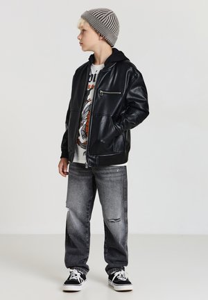 Giubbotto in similpelle nera con tasche frontali, t-shirt grafica bianca, jeans grigi strappati e berretto a righe grigio. Sneakers nere completano il look.
