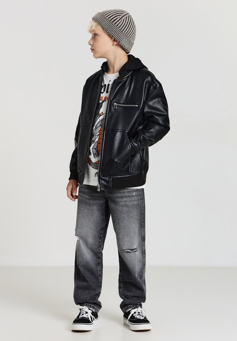 Giubbotto in similpelle nera con tasche frontali, t-shirt grafica bianca, jeans grigi strappati e berretto a righe grigio. Sneakers nere completano il look.