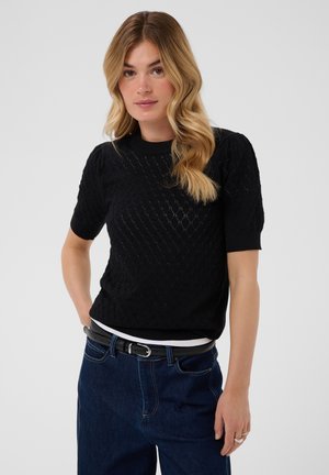 Femme aux cheveux blonds ondulés portant un haut noir à manches courtes en maille, un jean bleu foncé et une ceinture noire, debout devant un fond uni.
