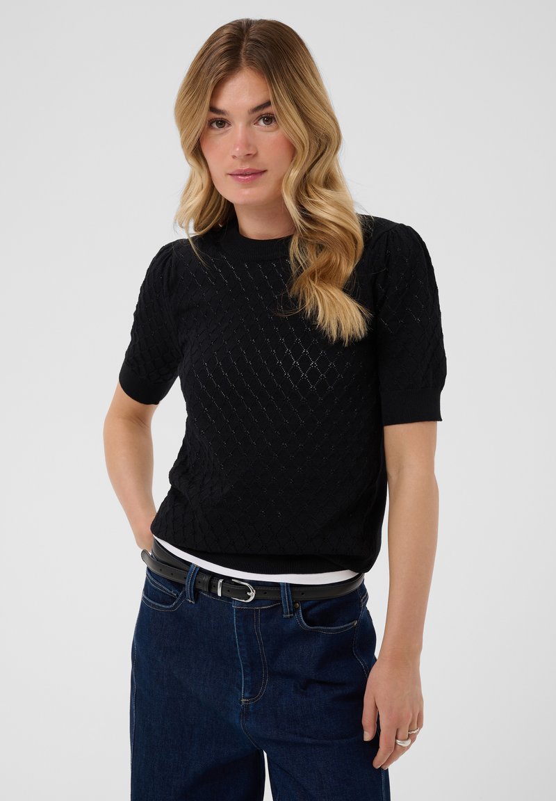 Femme aux cheveux blonds ondulés portant un haut noir à manches courtes en maille, un jean bleu foncé et une ceinture noire, debout devant un fond uni.