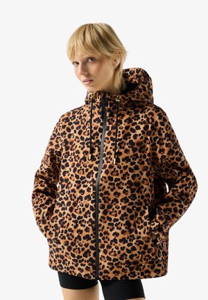 Nő barna és fekete leopárdmintás kapucnis dzsekiben, elöl cipzáras, fekete húzózsinórokkal, kezei a zsebekben, fehér háttér előtt.