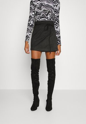 Gonna mini in suede nera con dettaglio in vita a lacci, abbinata a stivali alti fino alla coscia neri e a una maglietta a maniche lunghe con motivo bianco e nero.