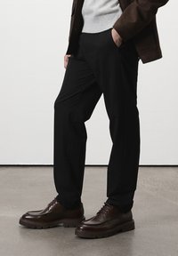 Homme portant un pantalon noir, des chaussures en cuir marron, un pull gris et une veste marron, se tenant avec une main dans la poche sur un sol gris.