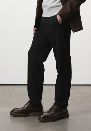Mann, der schwarze Hosen, braune Lederschuhe, einen grauen Pullover und eine braune Jacke trägt, steht mit einer Hand in der Tasche auf einem grauen Boden.