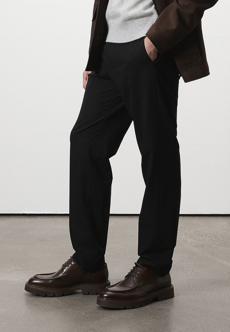 Homme portant un pantalon noir, des chaussures en cuir marron, un pull gris et une veste marron, se tenant avec une main dans la poche sur un sol gris.