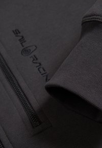 Schwarze Sweatjacke mit Reißverschluss aus strukturiertem Material, mit rundem Logo und dem Aufdruck "SAIL RACING" im Brustbereich.