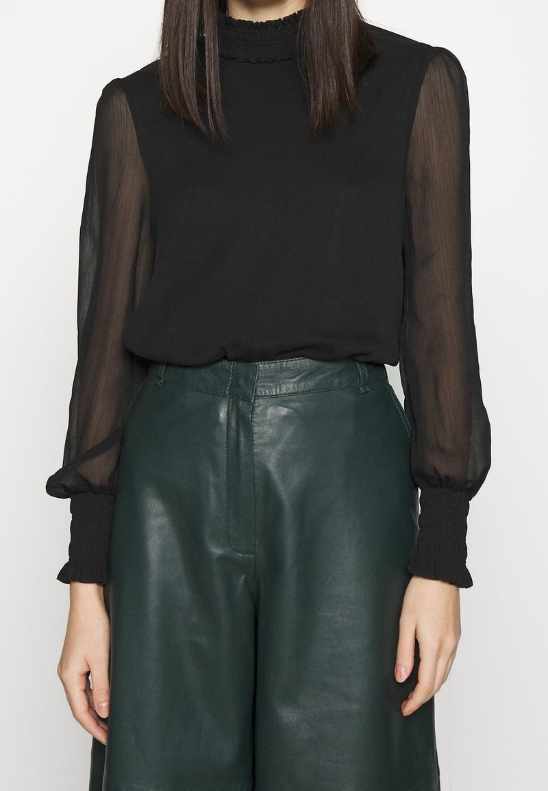 Femme portant un chemisier noir transparent avec des poignets à volants et un pantalon en cuir vert foncé taille haute, debout devant un fond uni.