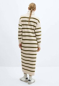 Langes, beige gestricktes Kleid mit dunkelgrünen horizontalen Streifen, mit hohem Kragen und lockeren Ärmeln, getragen mit hellen Socken und Schuhen.