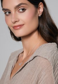 Srebrny naszyjnik o wzorze kwiatowym, noszony na beżowej, teksturowanej bluzce z kołnierzykiem. Włosy modelki są ciemne i falowane.