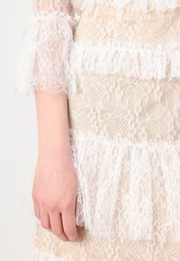 Rochie albă din dantelă cu volane suprapuse, având un material transparent, texturat și modele complicate; stratul de dedesubt de culoare deschisă este vizibil.