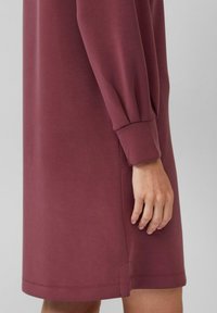 Bordeauxfarbenes Kleid mit langen Ärmeln, das eine weiche Textur und geraffte Ärmelbündchen aufweist. Der Saum ist leicht abgerundet und hat einen hohen Schlitz an der Seite.