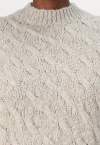 Kabelstrikket genser i lys grå farge, med høy hals og et teksturert mønster med hevede, sammenvevde design.