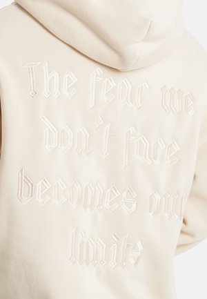 Beige huppari, jossa on huppu, ja jossa on teksturoitua valkoista brodyaattua tekstiä: "Pelko, jota emme kohtaa, muuttuu rajoiksemme," goottilaisessa fontissa.