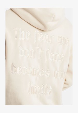 Beige Kapuzenpullover mit einer Kapuze, ausgestattet mit strukturiertem, weißem, gesticktem Text: "Die Angst, der wir uns nicht stellen, wird zu unseren Grenzen," in gotischer Schrift.