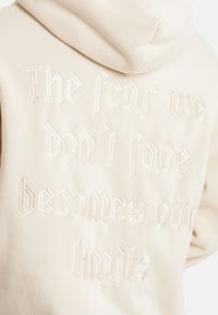 Béžová mikina s kapucí, s texturovaným bíle vyšitým nápisem: "Strach, kterému se nepostavíme, se stává našimi hranicemi," v gotickém písmu.