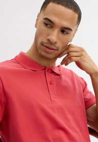 Polo shirt rosso realizzato in un tessuto liscio e texturizzato. Caratterizzato da un colletto, una chiusura a due bottoni e maniche corte. Primo piano sul colletto e sulla spalla del modello.