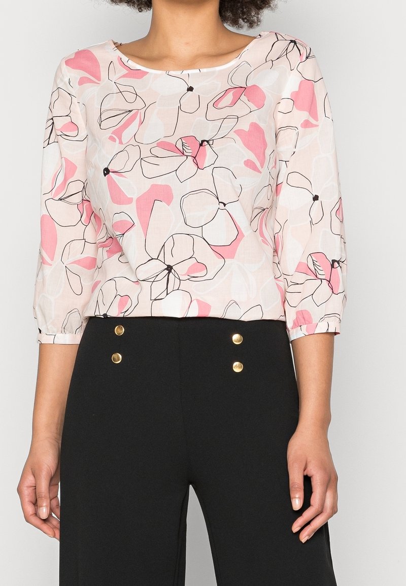 Femme portant un chemisier blanc avec un imprimé floral abstrait rose et noir et des manches trois-quarts, associé à un pantalon noir taille haute avec des boutons dorés.