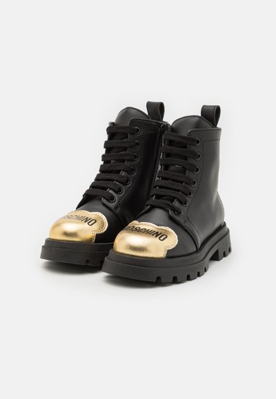 Botas de tobillo de cuero negro con una puntera dorada, suela de goma gruesa y cordones negros, con un logotipo impreso en la punta.