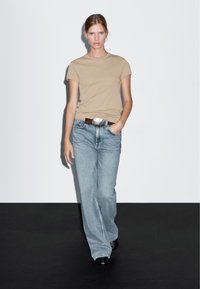 Béžové tričko s krátkým rukávem v kombinaci s světle modrými širokými džíny a hnědým páskem s velkou stříbrnou sponou. Jednoduché pozadí.