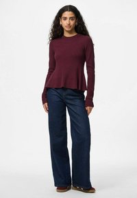 Sieviete ar krullainiem matiem, ģērbta bordo garā piedurkņu krekliņā, platajās tumši zilajās biksēs un bordo kurpēs, stāv uz viendabīga fona.