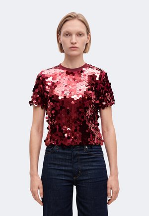 Un mannequin est de face, vêtu d’un haut rouge à manches courtes orné de sequins et d’un jean bleu foncé, sur un fond clair uni.