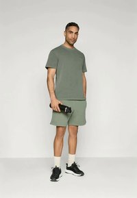 Meeste sportlik riietus rohelises: T-särk ja lühikesed püksid, puuvilla segu kangas, ümar kaelus, vabalt istuv, hoiab käes musta veepudelit, mustad tossud.