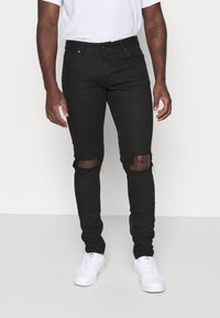 Svarta slim-fit jeans med rivna knän bärs av en person i en vit skjorta och vita Adidas-sneakers, som står mot en enfärgad bakgrund.