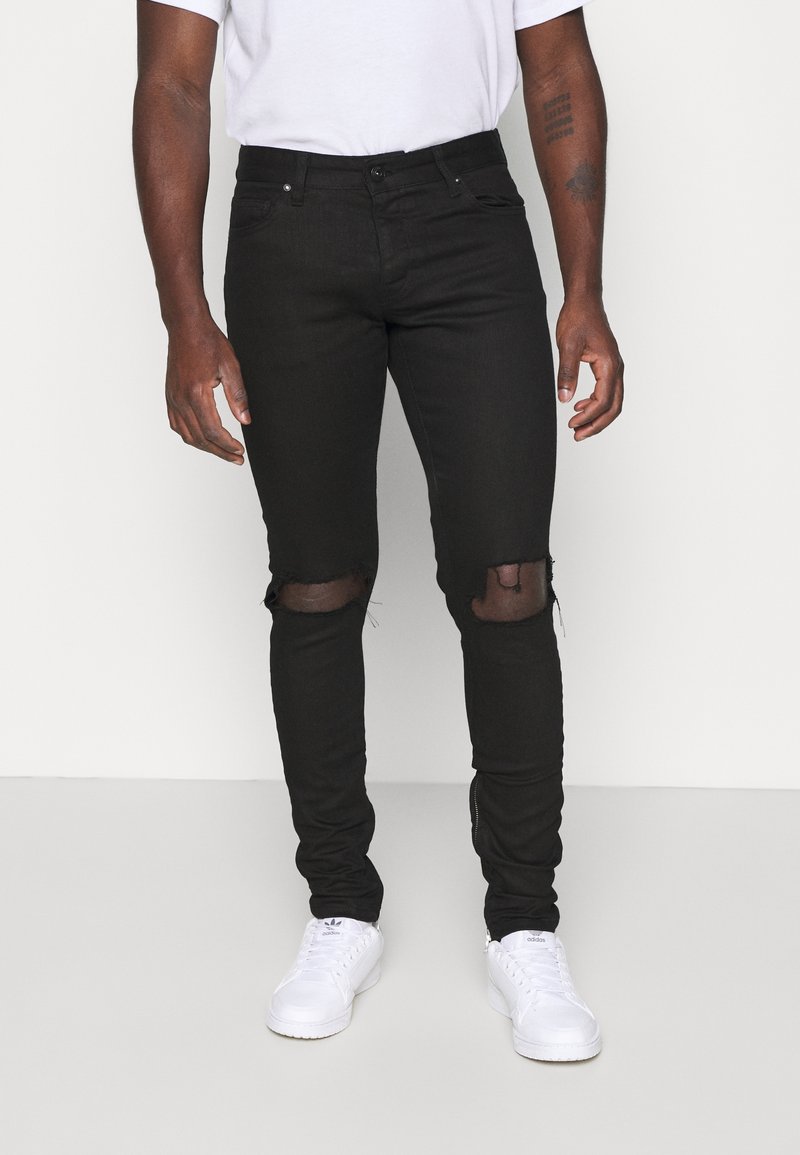 Svarta slim-fit jeans med rivna knän bärs av en person i en vit skjorta och vita Adidas-sneakers, som står mot en enfärgad bakgrund.