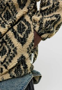 Beige und schwarze gemusterte Fleecejacke mit strukturierter Oberfläche, die ein "ORIGINALS"-Label trägt und über einem Denim-Kleidungsstück liegt.