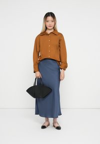 Ung kvinde iført en brun knap-op skjorte og en blå maxi nederdel, holder en sort geometrisk taske og har tofarvede flade sko på.