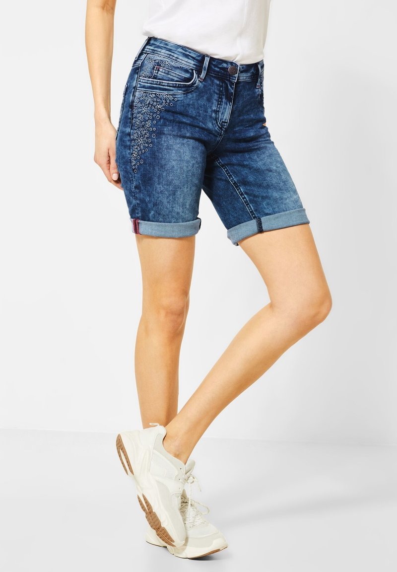 Mannequin portant un short en jean bleu foncé retroussé avec de petites décorations argentées sur la zone de la poche avant, assorti à des baskets blanches et un top blanc.