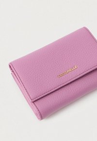 Folded textured light purple leather wallet with gold COCCINELLE logo on a white background.