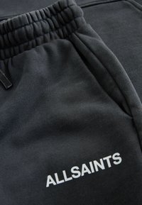Černé teplákové kalhoty s elastickým pasem, s jemnou strukturou a bílým logem "ALLSAINTS". Hladká látka s postranními kapsami.