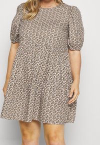 Kobieta w krótkiej sukience w kwiaty, o długości powyżej kolan, z okrągłym dekoltem i bufiastymi rękawami, stojąca na jednolitym tle.