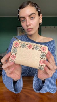 Cartera de cuero beige con acabado texturizado, que presenta un diseño floral geométrico en rojo, azul y negro alrededor de la zona del zipper.