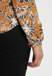 Bluzka w pomarańczowy wzór florystyczny z czarno-białymi detalami, dopasowane mankiety z guzikiem, zestawiona z czarnym materiałem.
