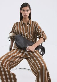 Joven mujer con camisa a rayas marrón y blanca y pantalones de pierna ancha sentada en una silla moderna sosteniendo un gran bolso de cuero negro.