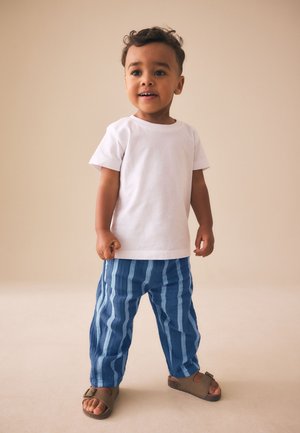 Mažas vaikas stovi, dėvėdamas baltą trumpomis rankovėmis marškinėlį, mėlynas dryžuotas kelnes ir rudus sandalinius, priešais paprastą smėlio spalvos foną.