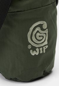 Zelena tkanina torba s teksturiranim površinom, s izvezenim spiralom logotipom u svijetlozelenoj boji i tekstom "WIP" sa strane.