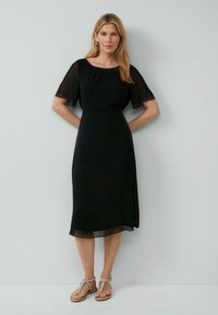 Abito nero con maniche corte trasparenti, scollatura arricciata e una silhouette fluida. Completato con sandali leggeri e con cinturini.