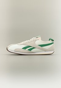 Λευκό και μπεζ αθλητικό παπούτσι με πράσινες ρίγες στο πλάι, λευκά κορδόνια και πράσινη θηλιά στη φτέρνα, τοποθετημένο σε ανοιχτό γκρι φόντο.