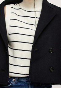 Giacca navy doppiopetto sopra un top bianco a coste con strisce nere, con collo alto e dettagli in zip argentata. Jeans in denim blu visibili.